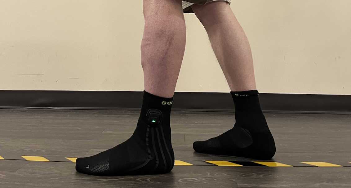 A person wearing black socks with built-in sensors stands on a wooden floor near yellow-and-black striped tape. The socks show a small green LED light on the side. Only the legs are visible.