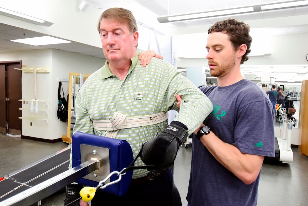 A physical therapist assists a man as he uses rehabilitation equipment in a therapy clinic. The therapist supports the man’s arm and back, guiding him through the exercise.