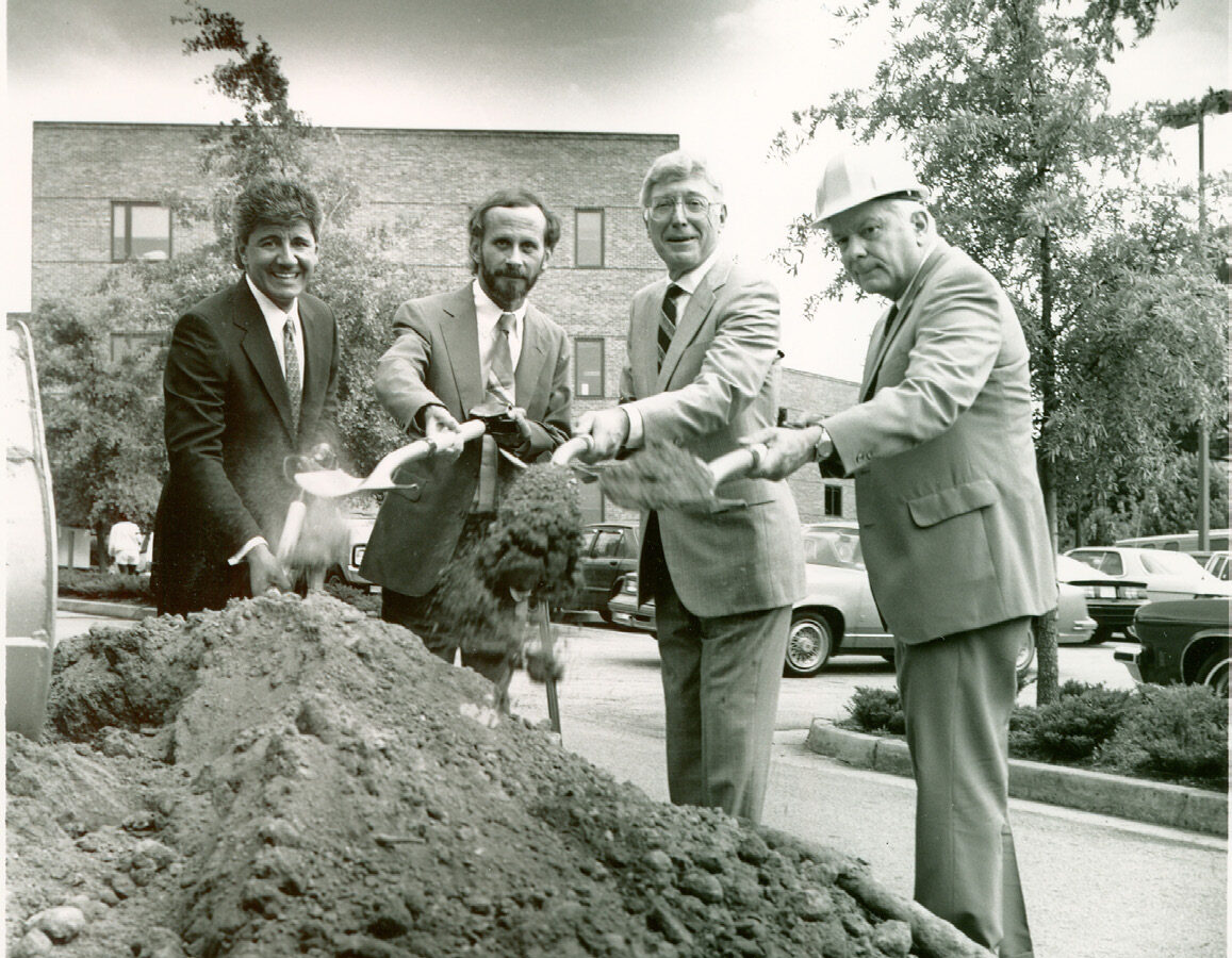 Four men in suits and a hard hat participate in a ceremonial groundbreaking, smiling while shoveling dirt. A building and parked cars are visible in the background.