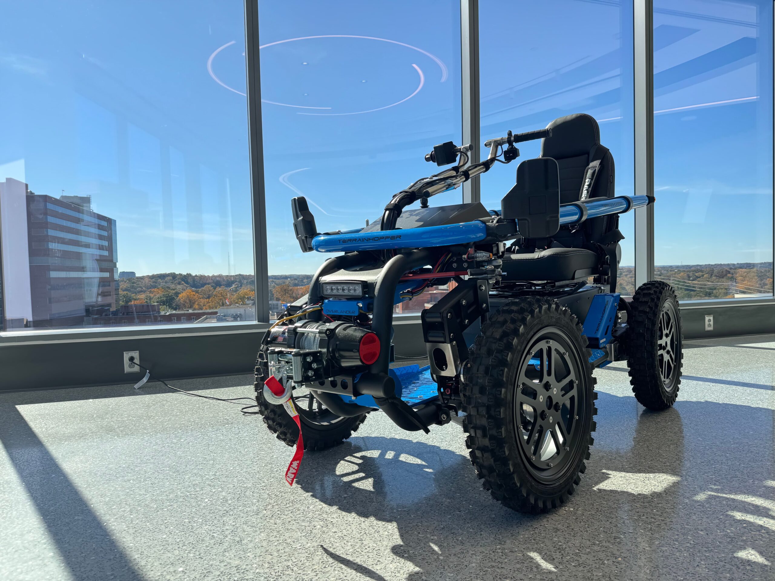 A rugged, all-terrain electric wheelchair with large off-road tires and a blue frame sits in a sunlit room with large windows overlooking a cityscape and trees.