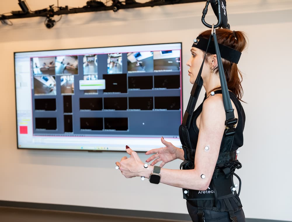 A person wearing a motion capture suit with sensors stands in front of a large screen displaying video feeds and data in a high-tech lab. The individual is focused, with hands raised in a gesture.